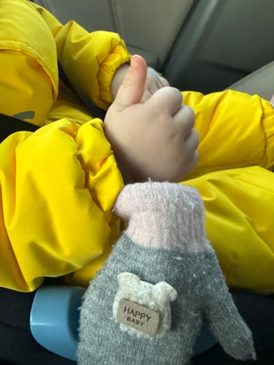 A close-up, downward-angled photograph depicts an indoor scene within the city of Bilimbay, Russia. A small, pale-skinned hand, presumably a child's, is positioned centrally, executing a 'thumbs-up' gesture with the thumb fully extended. This hand emerges from the cuff of a bright yellow, creased, and possibly padded jacket. In the lower right foreground rests a gray and light pink knitted mitten, exhibiting slight surface pilling. Attached to the mitten is a small, white, fuzzy, lamb-shaped appliqué with a tan rectangular tag beneath it, bearing the printed text "HAPPY BABY". A light blue, cylindrical object is partially visible in the lower left, adjacent to the yellow jacket. The background is blurred, showing muted dark gray and black tones consistent with an interior setting, possibly a vehicle passenger compartment.