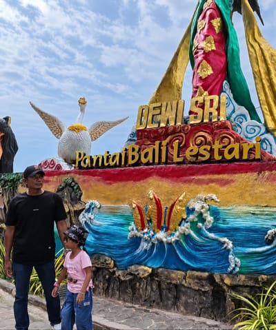 The image depicts a father and daughter standing in front of a colorful mural and statue, identified as being located at Pantai Bali Lestari in Kota Pari, Indonesia. The mural features a large, ornate statue of a figure, possibly Dewi Sri, adorned in red and green robes with gold accents. Below the statue, the words "DEWI SRI" are emblazoned in gold lettering, followed by "PantaiBali Lestari" in smaller gold letters. The mural itself depicts vibrant blue waves crashing against a rocky shore, with stylized golden fish or sea creatures emerging from the water. To the left of the main statue, a white swan-like sculpture with wings spread wide is visible. The father, wearing a black t-shirt, black cap, and blue jeans, stands to the left of the child. The daughter, dressed in a pink t-shirt and blue jeans, wears a patterned bandana on her head. They are both looking towards the mural and statue. The background shows a cloudy blue sky. The ground beneath them is paved with cobblestones.
