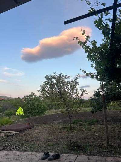 The image, possibly from Smilets, Bulgaria, shows a landscape under a sky with a large, elongated cloud tinged with a golden hue. The cloud extends vertically in the frame. The foreground features foliage and the frame of a structure, potentially a pergola, with vine leaves. In the background, there is a cultivated field with young trees. A reflective yellow safety vest hangs near the ground, possibly on a makeshift stand. A pair of shoes rests on a paved surface to the side.