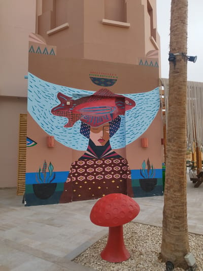 The image presents an outdoor scene, likely a public space, in Ras Abu Soma, Egypt. The focal point is a large mural on a building or wall, featuring a stylized depiction of a woman with a fish on her head, which rests in a hammock filled with water. The mural incorporates geometric shapes, patterned clothing, and plant motifs.
In the foreground, a red, artificial mushroom stands on a bed of small white stones. Beside it is a palm tree trunk with security lights attached. The ground is paved. A glimpse of outdoor seating can be seen behind the palm tree. The sky is overcast, suggesting diffused lighting conditions. No people are directly visible in the image, although the presence of seating and the artificial mushroom suggest a space intended for public enjoyment or decoration. The scene combines elements of art, nature, and urban design.
