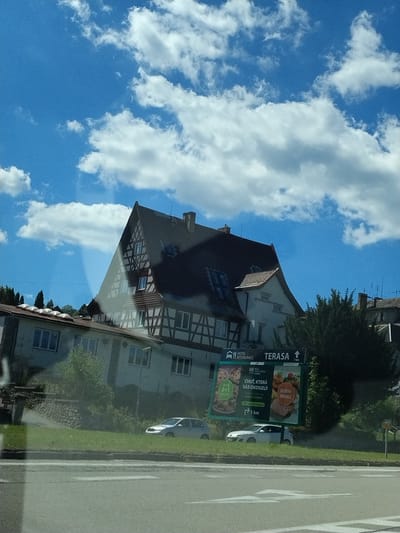 The image shows a view from a vehicle on a sunny day in Vimperk, Czechia. A traditional half-timbered building with a dark tiled roof dominates the right side of the frame. In front of it, a lighter colored building with a flat roof can be seen. A billboard for "HOTEL RESTAURACE TERASA" is prominently displayed, featuring images of food and indicating a distance of 1 km. A white sedan is parked on the grassy verge next to the billboard. The sky is blue with scattered white clouds. The foreground shows a road with painted white lines, indicating a pedestrian crossing. The overall scene depicts a roadside view in a town.