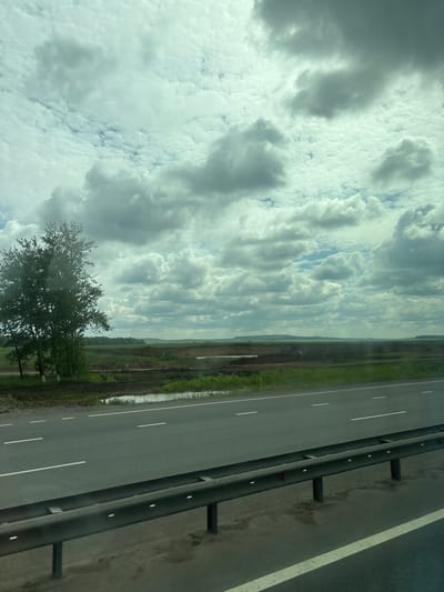 The image shows a highway with a metal guardrail in the foreground. The road has white lane markings. Beyond the road, there is a grassy field with some scattered trees and patches of water. In the distance, the landscape is mostly flat with rolling hills under a cloudy sky. There are no people or vehicles visible in the image. The scene appears to be in a rural or semi-rural area. The location of Poisevo, Russia is not visually identifiable from the image itself.
