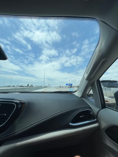 The image shows a view from the driver's seat of a car, looking out the windshield at a highway. The sky is blue with white clouds. The highway is clear, with light traffic. There are a few street lights visible. A sign for "American Airlines Arena" is visible on the right side of the highway. The car is parked near a large building, and there are trees and bushes in the distance. The image was taken in Westminster, California, at the coordinates 33.743, -118.003. A person's leg can be seen in the bottom right corner of the image. The car's interior is black leather. The image was taken during the day, under sunny conditions. The person is wearing a white shirt and blue jeans. The car is a modern vehicle with a sunroof. The highway is a four-lane road with a concrete barrier. The image is taken from a vehicle in motion, as the highway appears to be moving. The image is taken from a low angle, as the horizon is visible in the distance. The overall composition of the image is dynamic, with a sense of movement and motion. The subject of the image is the view from the driver's seat of a car, and the composition of the image reflects this. The image is well-composed and provides a clear and concise view of the scene. It is well-lit and the colors are vibrant. The image is sharp and clear.