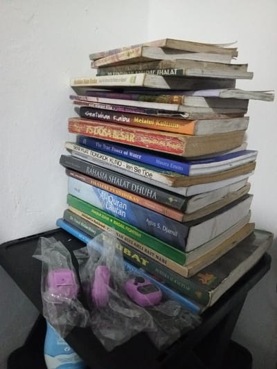 The scene is set indoors in the city of Al Haouafate, Morocco, against a plain, off-white wall. The primary subject is a tall, precariously stacked column of approximately 20-25 books, oriented with their spines facing outward. The books vary in size, thickness, and state of wear, with many appearing to be softcover and exhibiting signs of use. Several visible titles are in Indonesian or Malay, including "75 DOSA BESAR," "SENTUHAN KALBU Melalui Kultum," "The True Power of Water" by Masaru Emoto, "RAHASIA SHALAT DHUHA," "Al-Quran & Lautan" by Agus S. Djamil, and works by "Dr. Yusuf Al-Qardhawi."

At the base of the book stack, on a dark, flat surface that appears to be a table or shelf, are three distinct objects. Two small, clear plastic bags are present; one contains a light purple digital prayer bead counter (tasbih), and the other holds two small, light purple or pink plastic items, one of which has a hinged lid. Partially visible next to these items is the upper portion of a light blue bottle, possibly a personal care product, with the word "BABY" legible on its label. No individuals are present, and no actions or interactions are depicted within the frame.