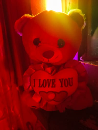 A close-up image shows a teddy bear lying on a surface. The teddy bear is predominantly orange, but the left side of its head is illuminated with a yellow light. Its face has black eyes and a dark nose. Next to the bear is a heart-shaped pillow bearing the words "I LOVE YOU" in black text. The background and surface appear red, possibly from lighting or the material of the surface. There is no information to suggest the image was taken in Okop, Bulgaria, and it's unlikely to determine the location of the image without more context.