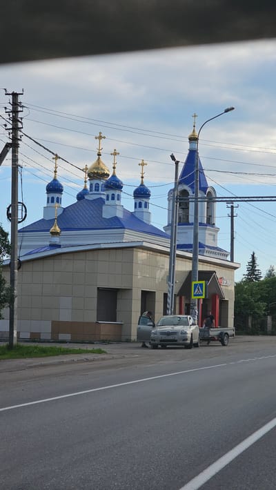 This is a medium-angle, eye-level, exterior, eye-level shot of a modern Russian Orthodox church with blue domes topped with golden crosses. The church is located in the city of Bolshebruzyanskoe, Russia. In front of the church, there is a light-colored building with beige tiled walls and dark-framed windows. Two people are visible outside the building. One person, wearing dark clothing, stands near the entrance of a silver car. The car is parked next to a small, single-axle trailer. Another person, wearing a light-colored jacket, is standing next to the driver's side door of the silver car. A pedestrian crossing sign is visible on a pole near the building's entrance. The scene is bordered by a road in the foreground, utility poles with many wires on the left side, and trees and a fence in the background. The sky is cloudy.