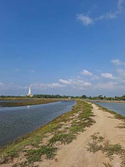 The image depicts an outdoor scene, likely in Dayahtuha, Indonesia, under a bright blue sky with scattered white clouds, suggesting a clear and sunny day. The perspective is vertical, rotated 90 degrees clockwise from a typical landscape orientation.
The primary subjects are a body of water, a dirt path, and vegetation. The water, appearing as a calm, reflective surface, occupies the right side of the image and stretches into the distance. Adjacent to the water is a narrow, sandy path, bordered by green grass and other low-lying vegetation. This path winds its way along the edge of the water, leading towards a distant point.
In the mid-ground, a tall, slender white structure resembling a minaret or tower stands prominently against the sky. It has a distinct, multi-tiered design. Beyond the minaret, a line of lush green trees forms a dense treeline, separating the water and path area from what might be land further beyond.
The scene appears tranquil and devoid of any human activity or visible text. The overall impression is one of a rural or semi-rural landscape, possibly near a body of water used for aquaculture or irrigation, given the structured appearance of the water channels. The time of day seems to be midday or early afternoon, judging by the brightness of the sky and the lighting on the landscape.