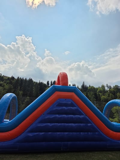 The image displays a close-up, low-angle view of a large, inflatable obstacle course on a grassy field. The inflatable structure is predominantly blue and orange, with large, curved blue sections on either side, and a central ramp-like structure with orange and blue accents. The texture of the inflatable material is visible, with stitching and air chambers apparent.

In the background, a dense forest of dark green evergreen trees covers a hillside. Above the trees, a bright blue sky is visible, dotted with fluffy white clouds and a patch of sunlit clouds in the upper left quadrant. The lighting suggests it is daytime, likely late morning or early afternoon.

The location is identified as the city of Neckertal, Switzerland. No people are present in the frame, and the focus is entirely on the inflatable structure and the natural environment. The image appears to be taken on a clear day with good visibility.