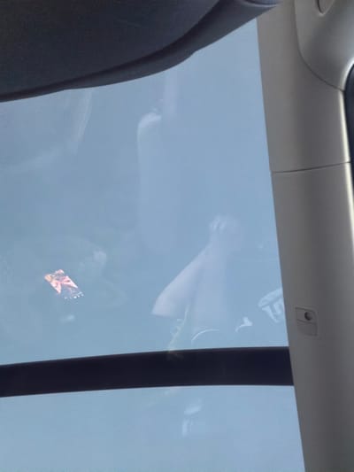 The image, taken in Dingle, Sweden, offers a view through a sunroof or panoramic roof of a vehicle. The sky appears overcast, casting a diffused light. A person is visible, their reflection or image subtly present on the glass. One hand is raised, and appears to be holding a smartphone. The phone screen displays a colorful image, possibly a photo or a graphic. The interior of the vehicle is also partially visible, including a dark trim along the top edge and a light-colored pillar on the right side, which has a small button or control on it. The overall impression is of someone using their phone while inside a vehicle, possibly on a journey.