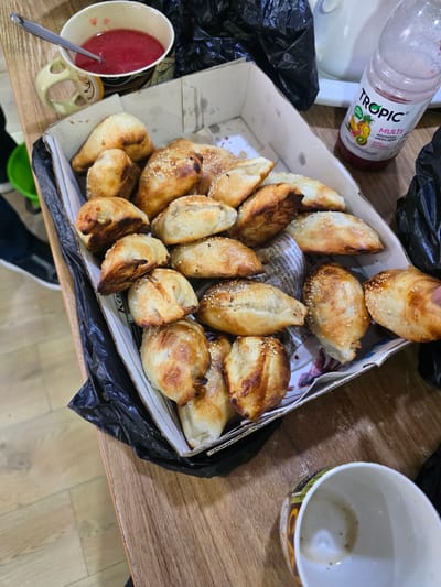 A close-up, eye-level, medium shot captures a tray of freshly baked, golden-brown pastries, sprinkled with sesame seeds, resting on a wooden table in what appears to be a casual setting, possibly a cafe or home in Karshi City, Uzbekistan. To the upper left, a mug filled with a red, pulpy beverage and a spoon sits beside the pastry tray. To the upper right, a clear plastic bottle with a pinkish-red liquid and a white label that reads "Tropic" is visible, alongside black plastic bags. The pastries, varied in shape and size, are arranged in a cardboard box lined with newspaper. The wooden table has a light, grainy texture. In the background, partially visible, is a white mug with a colorful, patterned rim, facing upwards. The lighting is natural and slightly diffused, highlighting the textures of the pastries and the table. No individuals are directly depicted in the frame, but the arrangement suggests a recent meal or snack. The overall scene evokes a sense of warmth and simple culinary enjoyment, characteristic of Uzbek hospitality.