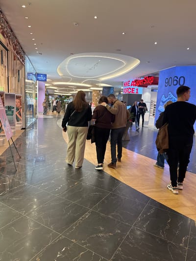 A busy shopping mall in Olginskaya, Russia, features a black and gold tiled floor, a bright white ceiling with recessed lighting, and various retail displays. In the foreground, a display case with cosmetic products is visible. Several people are walking or standing in the mall, some in groups, including a hugging couple. Various advertisements and signs are present.