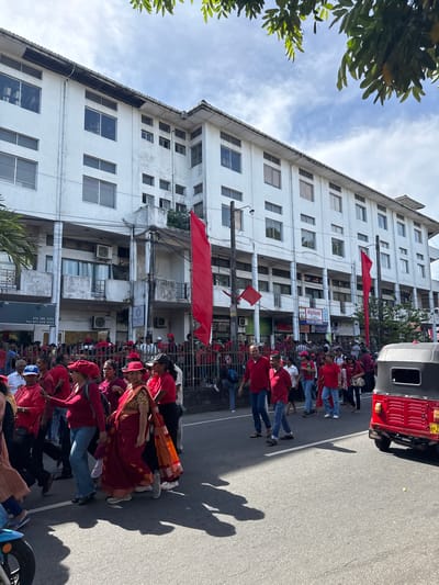 The image depicts a street scene in Delkanda, Sri Lanka. A group of people, many wearing red shirts and some red hats, are congregated on the road, likely for a protest or demonstration. Some women are wearing sarees, one with an orange accent. Red flags are hung from poles. A three-wheeled vehicle (a "tuk-tuk") is visible at the right edge of the image. Behind the crowd is a tall, white building with many windows. There are storefronts and businesses along the street. The signs for "Highland Milk Bar and Bakers," and "SENANAYAKE" are visible. Other signage includes telephone numbers: 074 382 1223 and Tel-011 478 3355. The sky is partly cloudy.