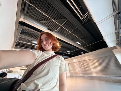 A low-angle shot shows a person with red hair, wearing a light-colored t-shirt with a brown shoulder strap, seemingly upside down in an industrial or commercial setting. The background consists of an exposed ceiling with a network of black beams, metal pipes, and some skylights or light panels. Parts of walls are visible, with one section featuring a horizontal, ridged texture. The location is unknown, but based on the instructions, it is believed to be in the city of Lautzenhausen, Germany.