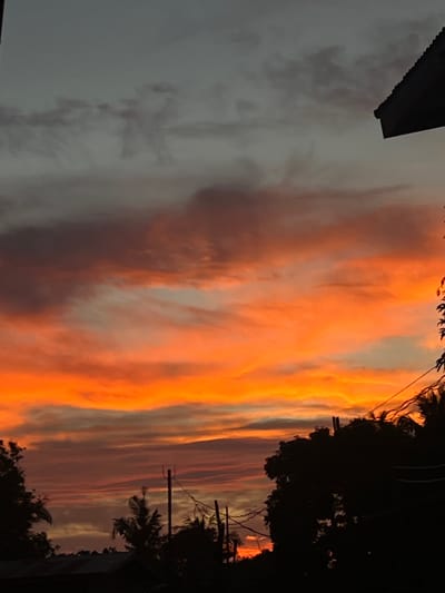 This image depicts a sunset in Cuyo, Philippines. The sky is filled with vibrant orange and red hues, transitioning to darker grays and blues at the top. Silhouetted against the colorful sky are trees and rooftops, suggesting a residential or semi-rural area. Utility poles and wires are also visible, further indicating a populated environment. The overall scene is one of natural beauty with elements of human habitation.