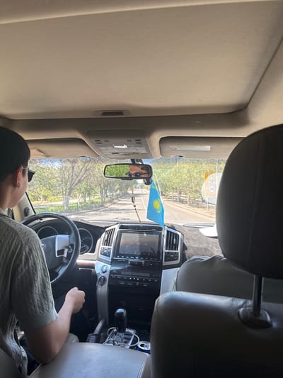 The image shows a view from inside a vehicle, looking forward. The driver, a young male wearing sunglasses and a light-colored sweater, is on the left. The front passenger seat is occupied by the back of another person's head, wearing a light-colored shirt. A Kazakhstan flag is visible on the dashboard. The vehicle is driving on a road lined with trees, suggesting a rural or suburban setting within or near Jezqazğan, Kazakhstan. A speed limit sign indicating 40 is visible on the right side of the road. The interior of the car includes the dashboard, steering wheel, gear shift, and a rearview mirror reflecting the driver. The overall lighting indicates it is daytime.
