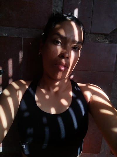 A person is lying on a brick surface, wearing a black sports bra. Sunlight filtered through a barred opening casts shadows across the subject's face, chest, and arms. The location is presumably outdoors, given the sunlight. The image orientation suggests the person is lying on their back, taking a selfie. The exact location within El Papelón, Venezuela cannot be determined from the image.