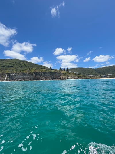 A coastline bisects the image. To the right, turquoise water reflects the bright sky, punctuated by white foam. To the left, a steep, verdant cliff face meets the water. Buildings are nestled within the vegetation near the cliff's edge. Above the cliff, a clear blue sky is scattered with puffy white clouds. The scene depicts a picturesque coastal landscape.