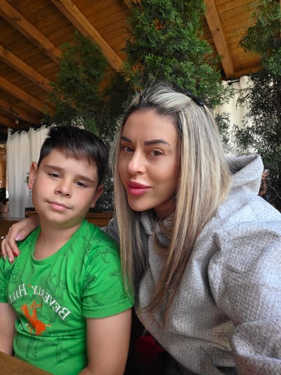 A selfie shows a woman and a young boy posing for a photo. The woman, on the right, has blonde hair and is wearing a gray hoodie. The boy, on the left, has short, dark hair and is wearing a green t-shirt with an orange horse graphic and white text. The woman's arm is around the boy's shoulders. They are seated outdoors under a wooden roof structure, with green foliage visible in the background. The scene suggests a casual outdoor setting, potentially a restaurant or cafe.