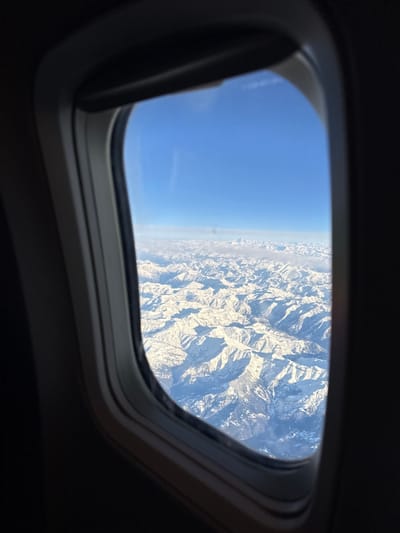 The image presents an aerial view through a rectangular aircraft window, observed from an airplane potentially in the vicinity of Colombier-Saugnieu, France. The foreground is dominated by the dark gray, slightly reflective frame of the airplane window, which borders a clear view of an expansive, snow-covered mountainous terrain under a bright, clear blue sky. Numerous rugged peaks and valleys are visible, entirely blanketed in white snow, with pronounced shadows cast by the sun, indicating direct overhead or high-angle illumination. A faint, distant line of haze or cloud formation is discernible above the mountain horizon, separating the snowscape from the unobstructed blue sky that fills the upper portion of the frame. No individuals, specific man-made objects within the landscape, or identifiable actions beyond passive observation are depicted. The environment is an alpine, high-altitude setting under clear weather conditions.