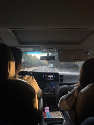 The image displays the interior of a vehicle, likely a minivan or SUV, viewed from a rear passenger's seat looking forward through the windshield. The scene is located on a multi-lane highway in San Miguel Topilejo, Mexico, during a period of low sun angle, indicating either late afternoon or early morning.

A driver, a person with short dark hair and glasses, is visible from the upper back and right shoulder, seated in the driver's seat with their right hand on the steering wheel. They are wearing a light-colored, possibly tan, short-sleeved shirt. A second individual, a front passenger with dark hair, is visible from the back of their head and upper torso, seated in the passenger seat and partially covered by a patterned blanket or shawl. Both occupants are illuminated by strong sunlight entering from the front and right side of the vehicle.

The vehicle's dark dashboard features a central infotainment screen displaying "18:03," "TuneIn," and specific music information. Below the screen are various climate and audio controls. The passenger-side sun visor is deployed. A sunroof is visible in the vehicle's ceiling. The center console between the front seats holds several objects, including a book titled "Jeffrey Archer - ONLY TIME WILL TELL," another book with a pink cover, and an electronic device connected by a pink cable.

Outside the vehicle, a paved highway with multiple lanes extends into the distance. Several vehicles, including passenger cars and a white semi-truck, are visible traveling in the same direction. A concrete barrier lines the left side of the highway, bordered by dense green trees. Similar vegetation flanks the right side of the road. The windshield exhibits minor reflections.