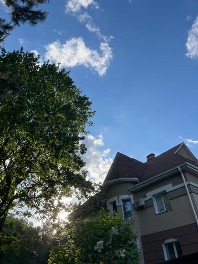The image is a low-angle shot of a house and trees against a blue sky with scattered white clouds. The house, located in Bogdanikha, Russia, has a brown shingled roof and beige exterior walls with dark brown trim. There are windows and an air conditioning unit visible on the side of the house. A large tree with green leaves occupies the lower portion of the frame, partially obscuring the sun, which creates a bright glare. Another, smaller tree is visible to the lower left. The sky is blue with scattered white clouds.