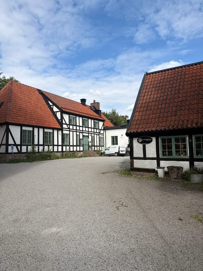 A wide-angle, eye-level shot depicts a gravel courtyard in front of a white and black timber-framed building with a red tile roof. The building features green-paned windows. To the right, another similar building is partially visible, also with a red tile roof and green window panes. A white van with "VÄRNAMO" text is parked in the background. The sky above is blue with scattered white clouds. The overall scene suggests a historical or rural setting. The building on the right has a sign that reads "LUNDABÖCKER CYKLAR & CAFÉ" and "Källängsvägen 11". This indicates the location is Lundaböcker, a shop and cafe specializing in books, bicycles, and coffee, situated at Källängsvägen 11 in Lund, Sweden.