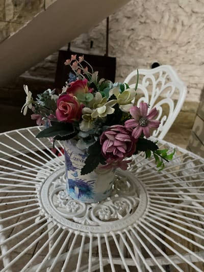 The image shows a close-up view of an artificial flower arrangement in a patterned vase. The vase is resting on a white, ornate, fan-shaped object, possibly a decorative screen or stand. The flowers are a mix of roses, daisies, and other filler flowers in shades of pink, white, and blue, with green leaves. The vase itself has a blue and white floral design, and a portion of text is visible, reading "Lava". The background appears to be a textured wall and possibly some wooden elements. The lighting suggests an indoor setting.