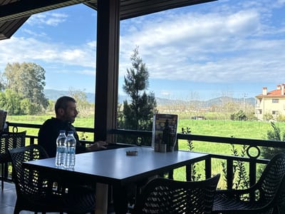 The image depicts an outdoor seating area of an establishment, likely a cafe or restaurant, located in Ula, Turkey. A single man, appearing to be middle-aged with short dark hair and a beard, is seated in profile at a dark rectangular table. He wears a dark long-sleeved shirt and faces right, looking out towards the panoramic view. Two clear plastic bottles of "Damla" brand water are placed on the table in front of him, along with a circular ashtray. A menu stand with a dark frame is also present on the table, to the left of the water bottles. The man's right hand rests on the table.

The seating area features dark, possibly wicker or plastic, chairs with patterned backs. The structure includes dark pillars and a dark railing with decorative elements, including a circular motif and leafy patterns. The ceiling of the structure is visible above.

Beyond the railing, the environment consists of a large, vibrant green field. In the mid-ground, various trees, some deciduous with bare branches and others with foliage, are visible. Distant mountains form the horizon under a bright blue sky with scattered white clouds. To the far right, a two-story building with a light-colored facade and a distinctive orange-tiled pitched roof is visible, suggesting residential or commercial architecture common in the region. Utility poles are faintly visible in the mid-ground between the field and the distant mountains.