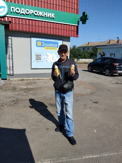 An elderly man wearing a black baseball cap, a dark blue jacket with blue stripes, and light blue jeans stands outdoors in front of a pharmacy in Chudniv, Ukraine. He is smiling and holding two ice cream cones, one in each hand. The pharmacy has a red tiled roof and a green sign with white lettering that reads "ПОДОРОЖНИК" (Podorozhnyk). A green cross-shaped pharmacy sign is attached to the building. A banner on the wall of the pharmacy advertises discounts of "up to 30%" and displays a QR code. A black car is parked behind the man to the right. The ground is paved with asphalt and gravel. The sky is clear and blue.