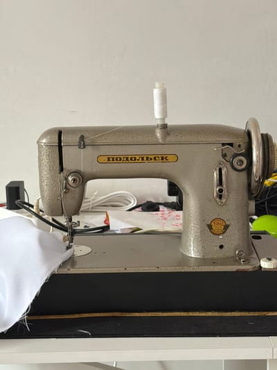 The image shows a vintage sewing machine, likely a "Podolsk" model, on a white table against a plain white wall. A spool of white thread is mounted on top of the machine. White fabric is positioned under the sewing needle, suggesting an ongoing or recent sewing operation. Various cords and other objects are visible in the background, partially obscured. The overall setting appears to be a domestic workspace. There is no indication of the city of Krasnyi Krym, Russia in the image itself.