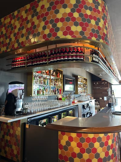 The image shows the interior of a bar or restaurant, potentially in Lake Buena Vista, United States, given the prompt's mention of the city. The scene is centered around a long bar with a striking design.
**Key Features:**
* **Bar Design:** The bar has a distinctive curved shape and is adorned with hexagonal tiles in a pattern of red, yellow, brown, and gray. This colorful tiling extends to the bar's facade.
* **Wine Display:** On one side of the bar, there's an elevated, conveyor-belt-like wine rack that stretches upwards. It's filled with rows of wine bottles, predominantly red wine.
* **Liquor Display:** Behind the bar are shelves displaying various liquor bottles.
* **Glassware:** Racks filled with wine glasses and other glassware line the back of the bar.
* **Equipment:** Standard bar equipment, such as coolers and ice bins, are visible behind the bar.
* **Lighting:** The lighting is bright, with natural light filtering in from a window above and artificial lights illuminating the bar area.
* **Overall Atmosphere:** The image suggests a stylish and well-stocked bar with a unique design, likely catering to a sophisticated clientele.
* **No People Present:** The image does not show any people in the scene.