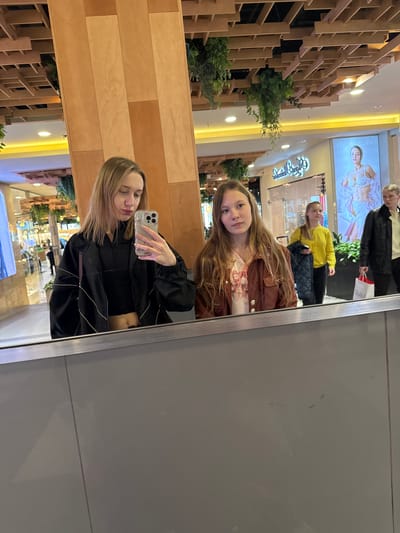 The image depicts a woman taking a selfie in a mirror located in a shopping mall, which can be identified as being in Ol'ginskaya, Russia, based on mall features in reflections. The mirror reflects herself and a younger girl behind her. The woman holds a smartphone and wears a black jacket over a cropped top. The girl behind her wears a brown jacket. Other shoppers are also visible in the background. Mall interior details, including ceiling lights, signs, decorations, and store displays, are visible in the mirror and the direct view of the scene.