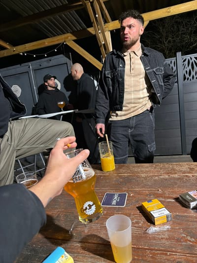 A group of people gather in an outdoor setting, possibly a patio, with a wooden table displaying drinks, cigarettes, and a coaster. A man in a denim jacket and checked shirt stands, while others sit. One person is pouring beer from a bottle into a glass. The area seems to be dimly lit, with wooden structures visible in the background.