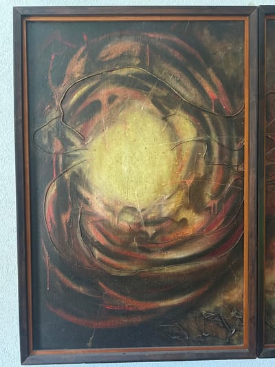 The image displays a framed abstract painting, part of a potential diptych or series, mounted on a plain, light-colored wall. The specific location within the city of Bogdanci, North Macedonia, cannot be determined from the visual information provided.
The central painting features a vibrant, luminous yellow-gold central area, appearing as a focal point or light source. This core is surrounded by turbulent, concentric swirling bands of dark red, orange, brown, and black, creating a sense of dynamic movement and depth. The artwork exhibits significant impasto, with thick applications of paint creating noticeable texture, particularly in the darker outer rings and some raised, winding lines in a lighter hue (possibly off-white or light brown) that traverse the composition. Visible drips of paint extend downwards from the brighter central area and some red elements. In the lower right quadrant, dark, textured, almost sculptural elements, resembling crumpled material, are integrated into the painting. The painting is enclosed by a dark brown wooden frame with a lighter inner edge. A portion of a second similarly framed artwork is visible on the right side of the image, suggesting continuity.