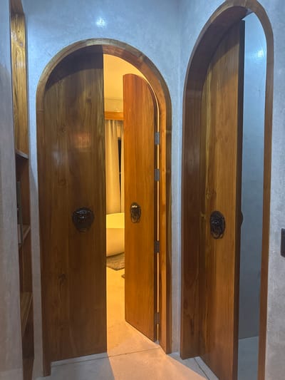 The image captures an interior hallway within a structure located in El Nido, Philippines. Two arched wooden doorways are prominently featured against light grey, textured wall surfaces. The wooden door on the left is partially open, hinged on its left, revealing a segment of an adjacent room. Within this room, a white freestanding bathtub is visible in front of light-colored curtains, illuminated by a warm, yellowish light. The floor of this visible room is light-toned. The wooden door on the right is closed or nearly closed. Both doors are equipped with dark, ornate, circular door knockers, resembling lion heads, positioned centrally at approximately waist height. Exposed metal hinges are visible on the left edge of the open door. The foreground floor is a light grey, possibly tiled or concrete, surface. A narrow vertical recess with wooden shelves is partially visible in the wall on the far left. No individuals or active interactions are depicted in the scene.
