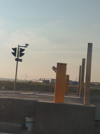A traffic light with a digital display showing "0 kg" is positioned on a pole in the foreground, to the left of the image. A security camera is mounted above the traffic light. To the right of the traffic light, a series of orange and beige pillars are spaced apart, suggesting a checkpoint or toll booth area. The ground in front of the pillars is paved, with some of it appearing wet. In the background, a low-lying building with a light-colored roof and blue accents is visible, along with scattered vegetation and a power line pylon. The sky is clear and pale blue, indicating daylight. A white bucket with a blue label is present in the lower left corner of the image, near a concrete barrier.
