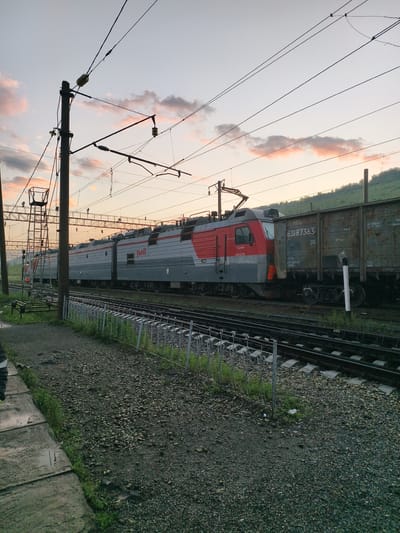 The image shows a freight train at dusk on a railway track in Obluchye, Russia. The train consists of a red and gray locomotive and several brown freight cars. The sky is a pale orange and pink, with scattered clouds. Overhead, there are power lines and a supporting pole. The foreground features a gravel trackbed, a concrete pathway, and a metal fence. The train is stationary.