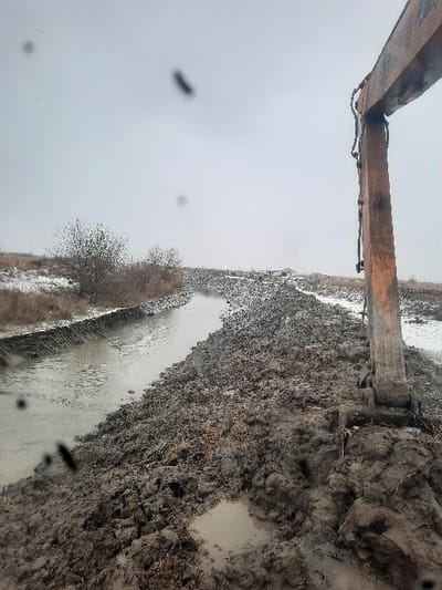 A muddy embankment borders a narrow stream or ditch, partially covered in snow. An orange excavator arm extends into the scene from the upper left. The sky is overcast and gray, with blurry spots on the viewing lens creating an obstructed view. Bare bushes line the edge of the water. The ground appears wet and churned, indicative of recent digging or excavation work.