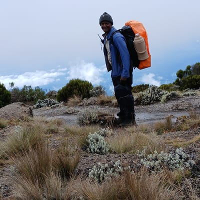 A single adult male is depicted standing on a rocky mountain path at Gilman's Point, Tanzania. The subject, of medium complexion, wears a dark grey knitted beanie, a blue outer jacket over a lighter shirt, dark trousers, and dark hiking boots. He is equipped with a large backpack covered by an orange rain cover, to which a light-colored, cylindrical flask is vertically strapped. The subject is oriented slightly to his left, facing the camera with a smile. The immediate foreground and mid-ground consist of sparse, clumpy yellowish-brown grass and small, silvery-grey, low-lying shrubs on dark, rocky terrain. A dirt trail is visible behind the subject. The background shows a vast expanse of white clouds extending below eye level, suggesting significant altitude, beneath a light grey, overcast sky.