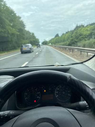 The image captures a first-person view from the driver's seat of a vehicle on a road in Stevenage, United Kingdom, during the daytime under an overcast sky. The road stretches into the distance, with two cars visible ahead in the same lane. Lush green trees and foliage line the left side of the road, with a guardrail separating the road from the natural embankment.
In the foreground, the right portion of a dark steering wheel, fitted with a textured, patterned cover, is prominent. To its left, part of the dashboard instrumentation is visible, including a speedometer marked with numbers from 0 to 120. A yellow warning light and a red warning light are illuminated on the dashboard. A control stalk, featuring symbols for various lights (including headlights and fog lights) and a "P" symbol, extends from behind the steering wheel. Additional discernible text on the gauges includes "H" and "C".
The perspective suggests the vehicle is either stationary or moving very slowly, as the speedometer needle is at zero. The other cars on the road also appear to be in a queue. The weather is cloudy, but there is ample daylight.
