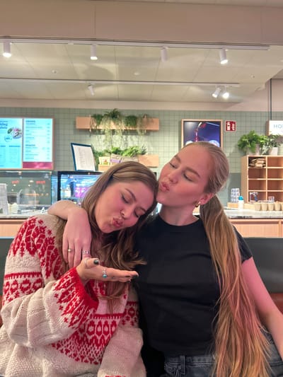 Two young adult females are seated side-by-side in a casual restaurant, identified as a Sickla Kebab establishment, located within the Westfield Mall of Scandinavia in Solna, Sweden. The female on the left, with light brown, wavy hair, wears a cream-colored sweater featuring a red fair isle pattern. She has multiple rings on her left hand, including one with a pearl-like stone, one with a turquoise stone, and several silver bands. Her left arm is draped over the shoulder of the female on the right. Her head is tilted, eyes closed, and lips pursed in a forward-facing kissing gesture. The female on the right has long, straight blonde hair styled in a ponytail over her left shoulder. She wears a plain black short-sleeved top and dark denim jeans, and a gold-toned hoop earring is visible on her right ear. Her eyes are also closed, and her lips are pursed, directed towards the left temple area of the other female. The background features a light green tiled wall adorned with wooden shelves holding various potted and hanging plants. To the left, a menu board displays "DRINKS" and "SNACKS" with prices, including "149:-" in Swedish Kronor. A display case is partially visible behind them. To the right, another section of wooden shelving holds multiple cups. A red fire extinguisher symbol is mounted on the green tiled wall. The ceiling is white with track lighting fixtures.