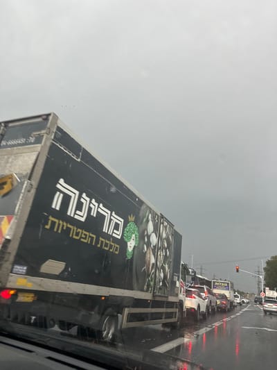 A black truck with Hebrew writing "מירקה מלכת הפטריות" (Mirka, Queen of Mushrooms) and an image of mushrooms is on the road. The truck's rear depicts more mushroom images. Traffic is heavy with multiple cars lined up ahead. The sky is overcast and gray, suggesting a rainy day. The scene is viewed from inside a vehicle, likely stuck in the traffic.