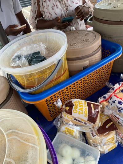 The image displays a close-up view of a food vendor's stall or market stand in Igando Oloja, Nigeria. The foreground features a blue surface upon which various food items and containers are arranged. A clear plastic bucket, approximately half-full of a yellow-orange liquid, likely soup or stew, is prominently centered, containing a clear plastic bag filled with multiple green utensils. Adjacent to this bucket is an orange plastic basket with a blue rim. Several beige, insulated food containers are present, with one larger container displaying the partially visible text "WANWE" on its side. Below the basket, multiple loaves of "Good Taste Butter Bread" are visible, individually packaged in clear plastic. In the bottom-center, a clear rectangular plastic container holds several white, spherical objects, consistent with boiled eggs. Two individuals are partially visible in the background. On the left, an individual wearing a white shirt holds a white smartphone in their left hand. On the right, another individual, wearing a light-colored blouse with a subtle repeating pattern, holds a teal-colored smartphone in their right hand. Their left hand is also visible near the center. The scene captures an active commercial food environment.