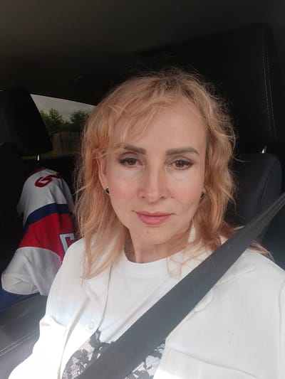 A woman with blonde, shoulder-length hair and light skin is seated in the driver's seat of a car, wearing a white blazer over a white t-shirt. A black seatbelt is visible across her chest. In the background, a red, white, and blue sports jersey with the number "6" is hanging from the headrest. The car appears to be parked outdoors, with a fence and trees visible through the car window. The setting is the city of Kuzhendeewo, Russia.