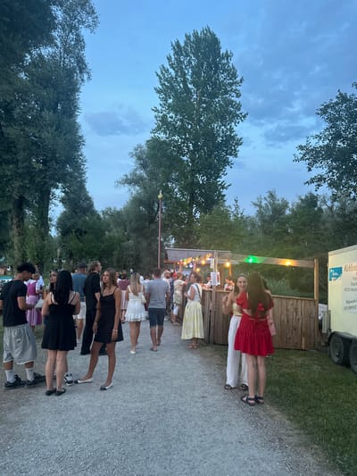The image depicts an outdoor event in the city of Rimsting, Germany, likely a festival or fair, with many people gathered. The scene is set with a twilight sky overhead, characterized by scattered clouds and a soft blue hue, bordered by tall, leafy trees on the left and right sides of the frame. In the background, a wooden structure resembling a food or drink stand is visible, adorned with string lights that are illuminated. A white trailer with signage is parked to the right.
A diverse crowd of individuals, dressed in casual and festive attire, are milling about on a gravel path. Several people are engaged in conversation, while others appear to be queuing at the stand. The individuals are of various ages and genders, and their interactions suggest a relaxed and social atmosphere. The ground is covered with gravel, with patches of green grass visible on the sides. The overall environment suggests a pleasant evening gathering in a park-like setting.