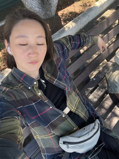 The image shows a close-up, high-angle shot of a person sitting on a wooden park bench. The person, who appears to be a young woman, is wearing a plaid flannel shirt over a black top, a silver necklace with pearl-like pendants, white wireless earbuds, and a grey fanny pack. Sunglasses are placed on the bench next to her. Her eyes are closed and her face is angled slightly downwards. The bench is made of dark wooden slats and is situated outdoors. In the background, there is a concrete curb and the base of a tree with visible bark. The ground is covered with brown dirt and some scattered pinecones. Sunlight is casting dappled shadows across the bench and ground, suggesting it is a sunny day. The overall scene is relaxed and casual. There is no information in the image to suggest the location is Pereyaslavka, Russia.