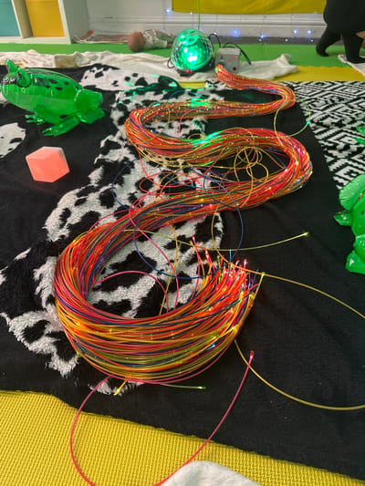 The image depicts a sensory play setup on a black rug with a cow print pattern and a yellow textured mat. In the foreground, a large bundle of colorful fiber optic cables, illuminated with small lights, is coiled. Individual strands extend outwards, some with glowing tips. To the left, a translucent green inflatable frog toy and a pink illuminated cube are visible. In the background, a green disco ball light and a small electronic device are present. A person's legs, wearing black tights and shoes, are visible on the right side of the frame. The scene appears to be indoors, possibly in a sensory room or playroom, but no specific location in Wakefield, United Kingdom can be identified from the visual information provided.