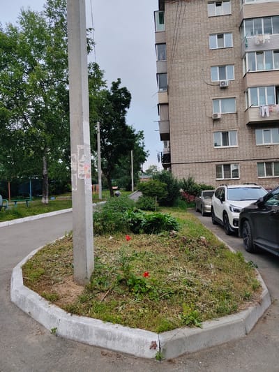 The image depicts an exterior urban scene in Trudovoye, Russia, featuring a multi-story brick apartment building situated to the right. The building has a facade constructed of light-colored bricks with multiple windows and balconies. Clothes are visible hanging from a balcony. In front of the building, several cars are parked along a paved area, including a white SUV and a dark SUV, with a lighter-colored sedan parked further back. To the left of the building and cars, there is a concrete utility pole with some torn posters attached to it. Adjacent to the pole is a small, irregularly shaped patch of dry grass and some scattered green vegetation, bordered by a concrete curb. Beyond this patch, a paved pathway curves to the left, leading towards a green area with trees and what appears to be playground equipment or park benches in the background. The sky is overcast.
