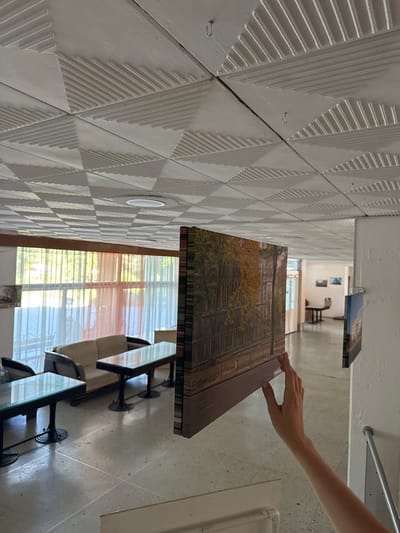 A person's hand with manicured fingernails is reaching out to touch a framed piece of art. The artwork depicts a street scene with trees in autumn foliage and architectural elements. The art is displayed in what appears to be a gallery or exhibition space, as indicated by the consistent wall treatment and other artworks visible in the background. The walls are decorated with a textured pattern of raised geometric shapes, resembling a grid of inverted triangles. There are also framed pictures hanging on the far wall, and a table with chairs is visible. Natural light streams in from a window covered with blinds, suggesting it is daytime. The overall impression is one of a calm, art-focused environment.