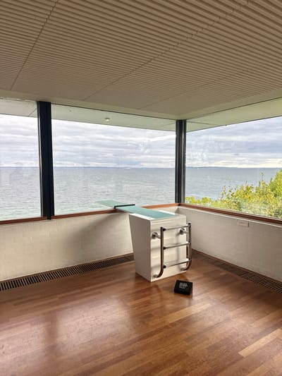 The image depicts an interior scene at the Louisiana Museum of Modern Art in Humlebæk, Denmark. The room features a polished light brown hardwood floor, white painted brick walls below expansive, black-framed windows, and a light-colored, horizontally slatted ceiling. Beyond the windows, the grey, choppy waters of the Øresund are visible under a cloudy sky, with a distant landmass on the horizon and faint indications of small vessels. Sparse green foliage is present outside the lower right window section.
Centrally positioned on the hardwood floor is a white, freestanding art installation resembling a diving board structure. This piece comprises a rectangular base featuring three curved, polished metal horizontal bars on its front face. A light green-surfaced diving board extends horizontally from the top of this base towards the windows. Directly in front of the installation, a small, black rectangular sign with white text, likely stating "PLEASE DO NOT TOUCH," rests on the floor. Horizontal gridded floor vents are visible along the base of the windows. No individuals are present in the scene.
